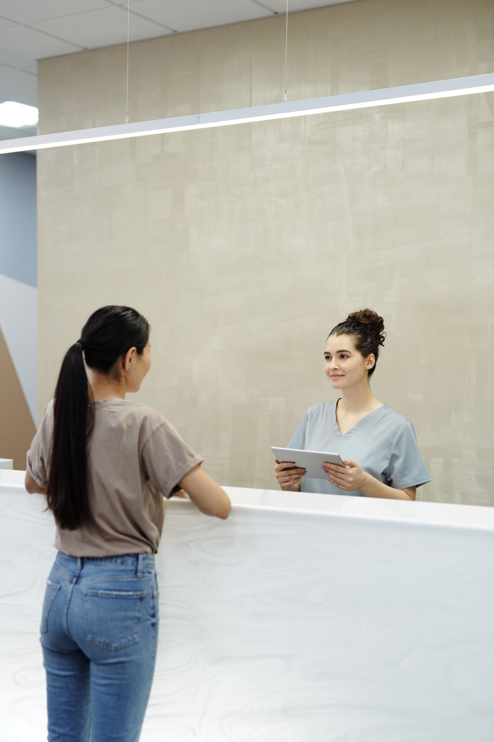 Profissional de saúde com tablet atendendo uma visitante vista de costas na recepção; fotografia de banco.