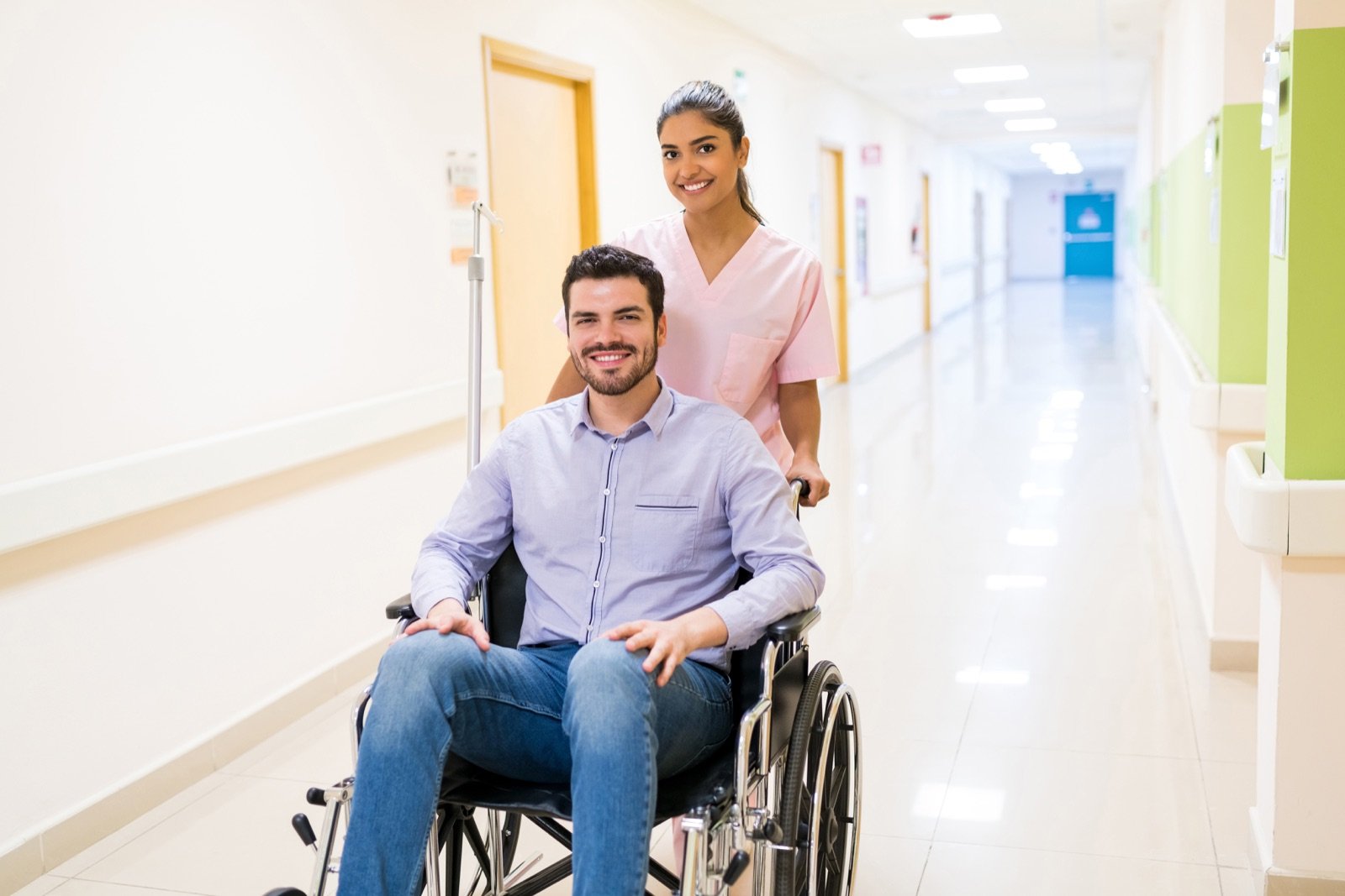 Pessoa em cadeira de rodas acompanhada por profissional de saúde, ambos sorrindo em corredor claro; fotografia de banco.