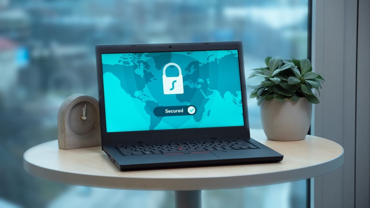 Laptop sobre mesa redonda exibindo um ícone de cadeado, ao lado de um vaso de planta e de um relógio; fotografia de banco.
